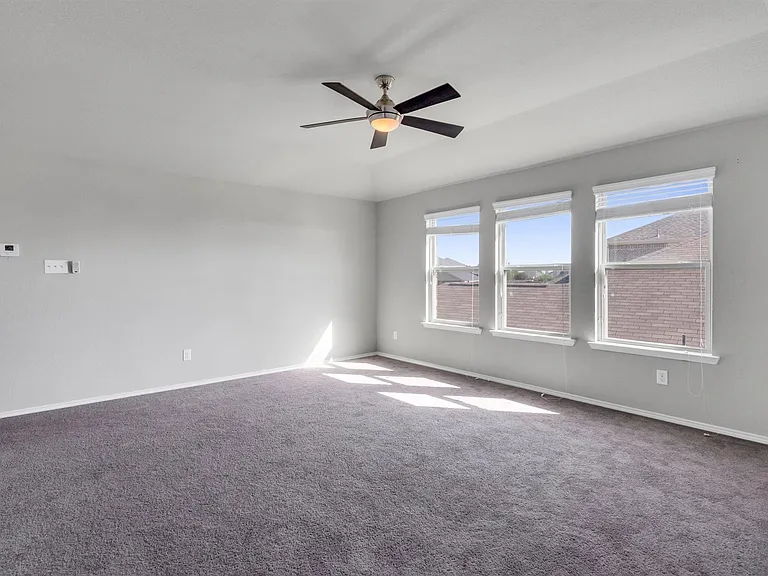 Spacious room featuring gray carpeting, multiple windows with blinds, a ceiling fan with integrated lighting, and a tray ceiling detail