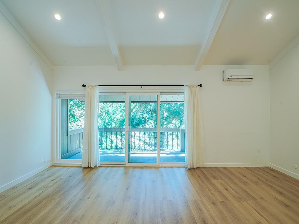 View of large living room from the fireplace wall looking out toward the balcony and tree tops. New HVAC split unit with remote control can be seen top left corner.