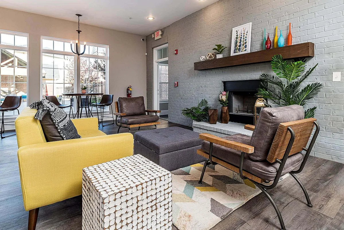 Inviting living room with modern decor, featuring a cozy fireplace, vibrant yellow sofa, stylish leather chairs, and natural light from large windows. - The Grand at Shoaff Park