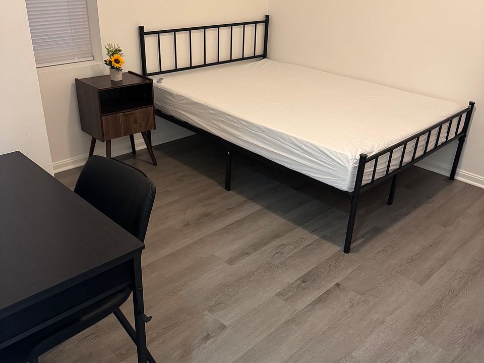 Furnished bedroom with a full-size bed, nightstand, and dedicated desk setup, plus a window with blinds and wood-look floors.