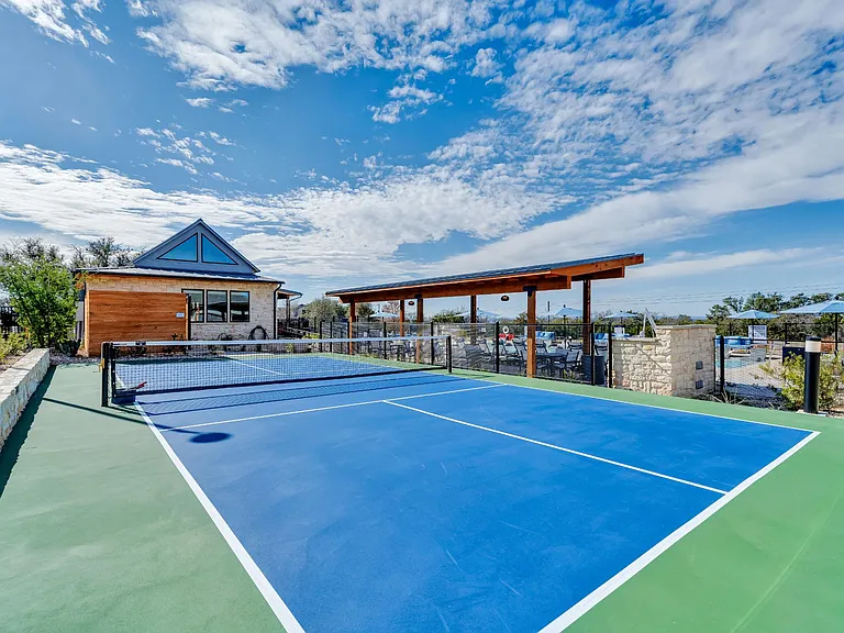 A tennis court with a blue surface and white lines, surrounded by a fence and a building with a blue roof.