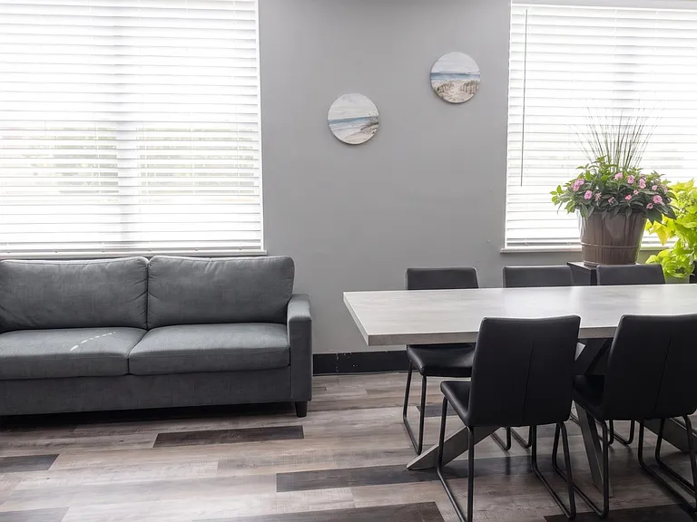 Open common area with a dining table, seating for six, and a sofa, plus large windows with blinds and durable flooring.