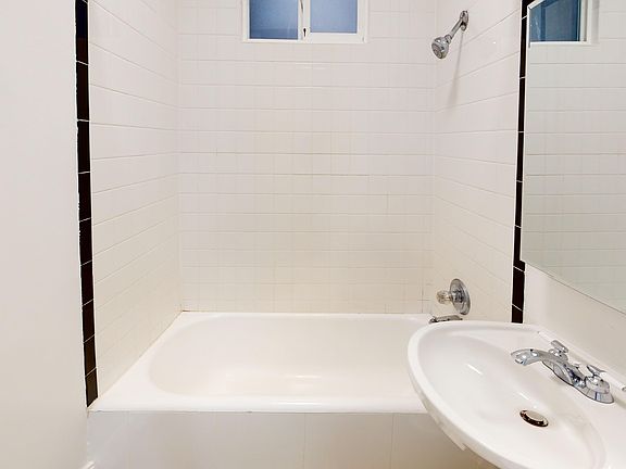 A clean, small bathroom features a bathtub with a showerhead, a sink with a chrome faucet, and a large mirror. The walls are tiled, conveying a simple, minimalist style.