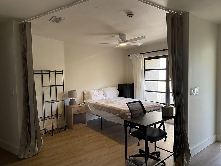 Private furnished bedroom featuring a bed, desk workspace, open shelving, and a large window with curtains and ceiling fan.