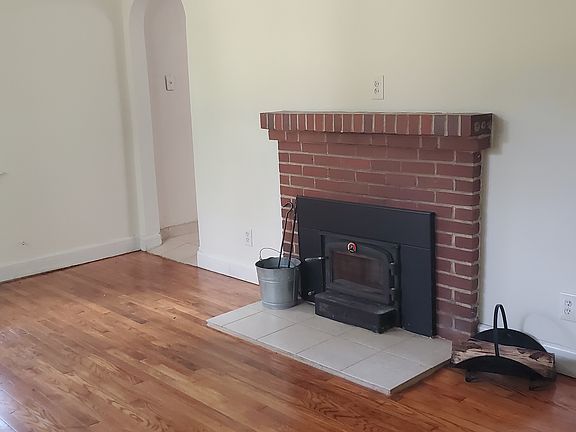 Living room showing hard wood floor, brick fireplace, fireplace insert with blower, wood rack, ash bucket and fireplace utensils, all of which come with the house.