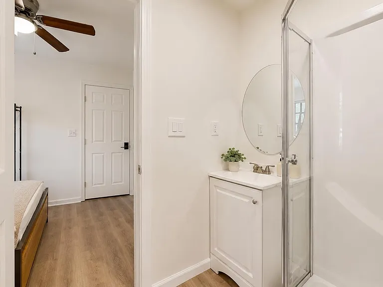 Clean bathroom with a glass-enclosed shower, single-sink vanity, and round mirror, plus easy access from the hallway.