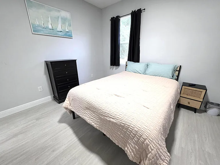 Furnished bedroom with a full-size bed, nightstand, and dresser, plus a window with curtains for privacy and light control.