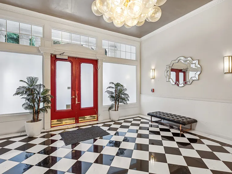 Grand entrance to a building, featuring a double door with a handle, a chandelier hangs from the ceiling, and two plotted plants, one on each side of the door