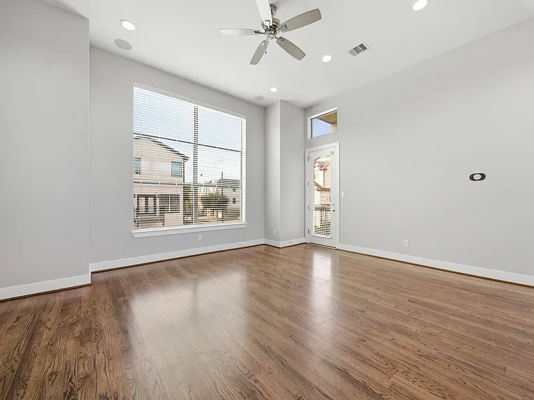 Bright and spacious living area filled with natural light, showcasing rich hardwood floors, neutral tones, and a modern ceiling fan. Expansive windows and a glass door create a seamless indoor-outdoor connection.