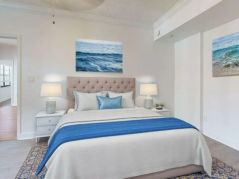 Charming bedroom with elegant decor, featuring ocean-themed art, a cozy bed with a blue accent pillow, and calming lamps on modern nightstands.