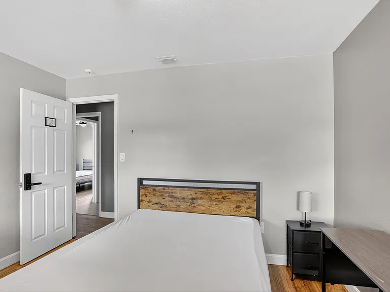 Furnished bedroom with a bed, nightstand, and work desk, plus neutral finishes and easy access to the hallway.