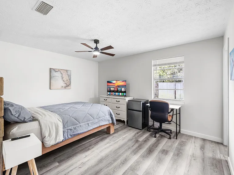 Furnished bedroom with ceiling fan, window, work desk, dresser, and in-room mini fridge for added convenience.