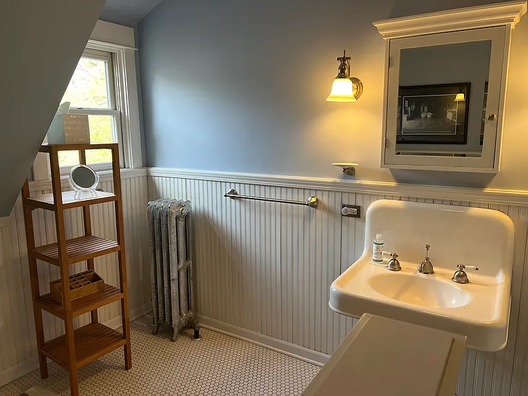 Front bathroom is spacious and sunfilled with wainscoating drawing on traditional theme: hexagonal ceramic tile floor, traditional medicine cabinet, antique wall hung porcelain sink, solid caste iron clawfoot tub and linen closet