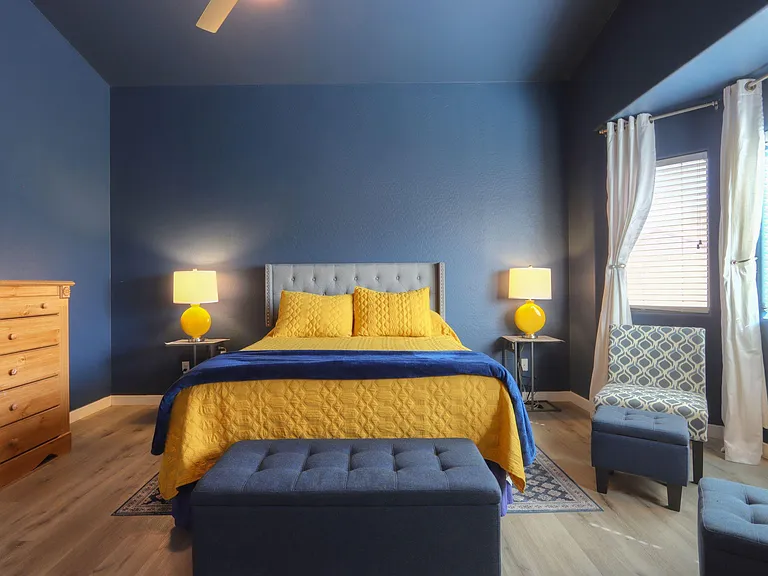 Spacious bedroom with a full bed, dresser, reading chair, and large windows with curtains for natural light and privacy.