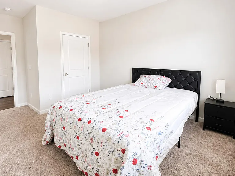 Furnished bedroom with a full bed, nightstand and lamp, plus a large window with blinds and neutral finishes.