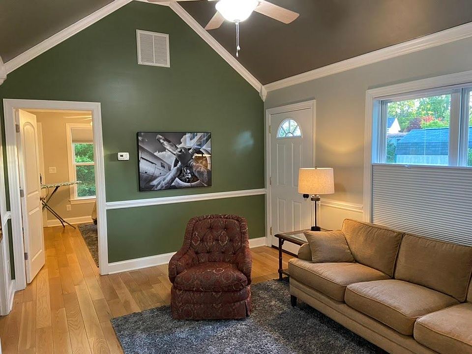 Living room has vaulted ceiling, ceiling fan, top-down/bottom-up cellular solar shades on west-facing windows, couch, chair, cocktail tables, area rug, and wall-mounted flat screen TV