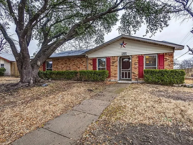 Brick facade residence featuring a prominent mature tree, red window shutters, and a concrete pathway