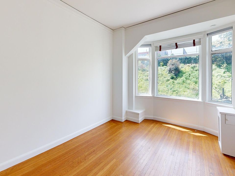 Bright, empty room with hardwood floors and large windows, featuring a scenic view of greenery outside