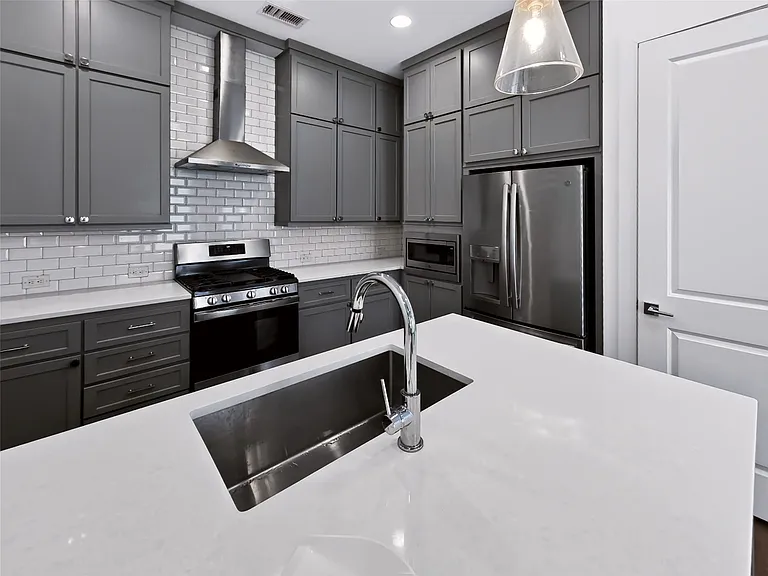 Modern kitchen featuring a large island with an undermount sink and chrome faucet, sleek grey cabinetry, a subway tile backsplash, stainless steel appliances, and white countertops
