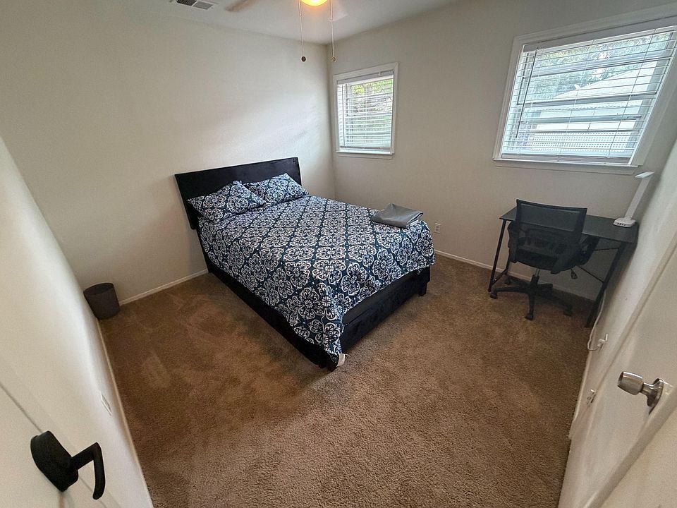 Private carpeted bedroom with a full bed, two windows with blinds, ceiling fan, and a dedicated desk for work or study.