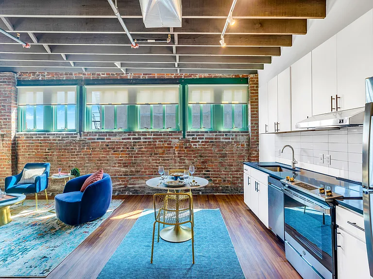 Urban Industrial Living: A spacious kitchen and living area framed by exposed brickwork, rustic wooden ceiling beams, and large windows, accentuated by pops of blue from the furniture.