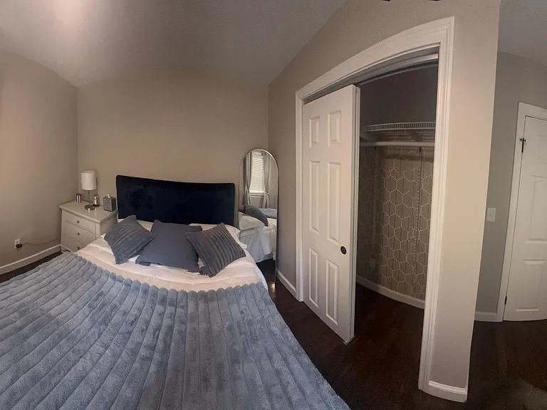 Furnished bedroom with dark wood floors, two windows with blinds, and a closet with built-in shelving for storage.