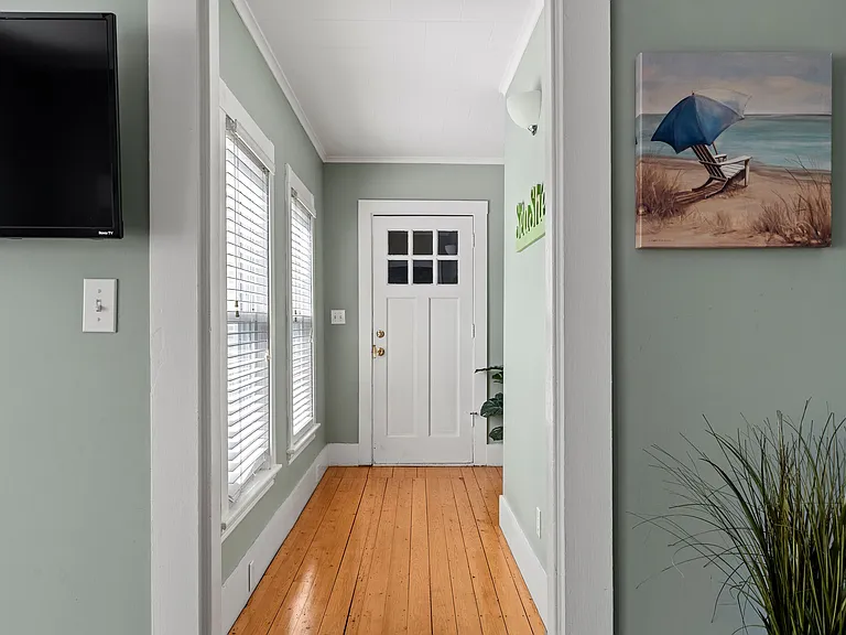 Welcome home! This charming entry area features a classic white door with window panes and original hardwood floors. The walls are a soothing sage green, complemented by nautical/beach-themed decor ("Sea Shells" sign) and bright natural light from the sid