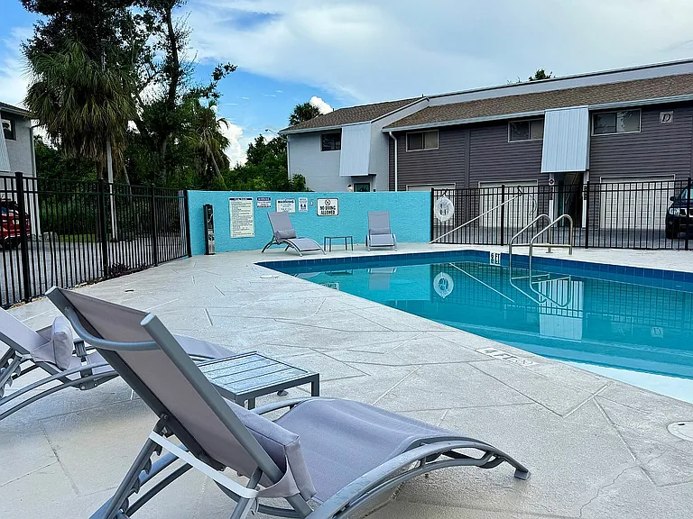 Inviting poolside area with modern lounge chairs and a serene blue swimming pool, surrounded by lush greenery and contemporary apartments.