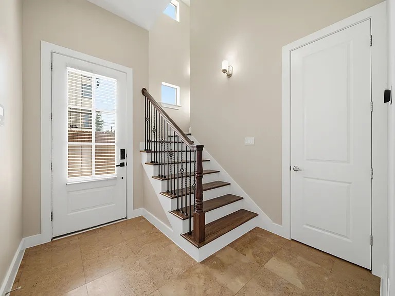 Bright, inviting entryway featuring a warm wood staircase with elegant iron railings and a refined neutral palette. Natural light flows through the space, enhanced by a stylish wall sconce, creating a welcoming and sophisticated first impression.