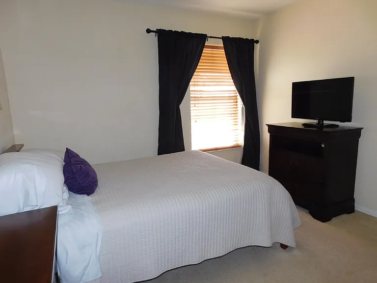 Furnished bedroom with a comfortable bed, window with blinds and drapes, and a dresser topped with a TV for easy lounging.