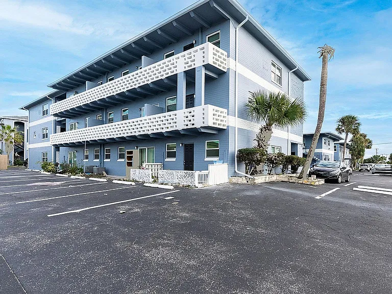 Charming coastal apartment complex with blue exterior, ample parking, and lush palm trees, perfect for serene living.