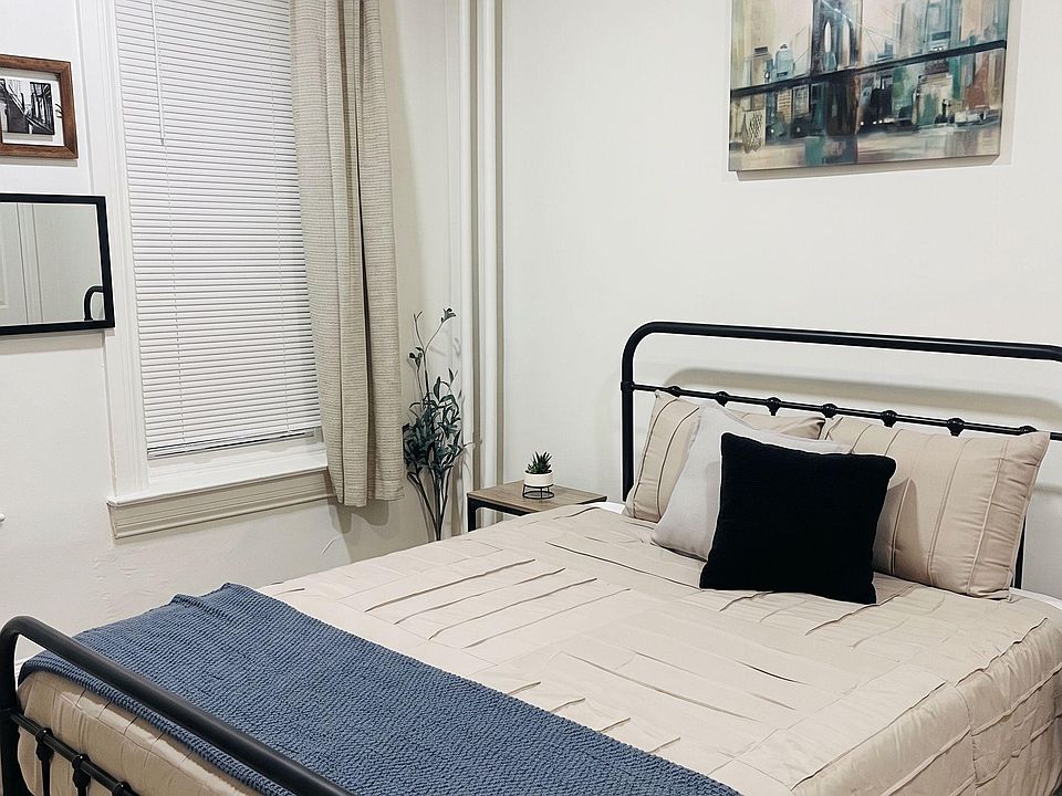 Furnished bedroom with a full-size bed, side table, and window with blinds and curtains for light control.
