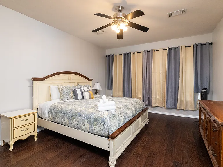 Relax in the primary bedroom with a king bed, soft bedding, TV, and warm lighting. Features a ceiling fan, dresser, room-darkening curtains, and a private attached bathroom for added comfort.