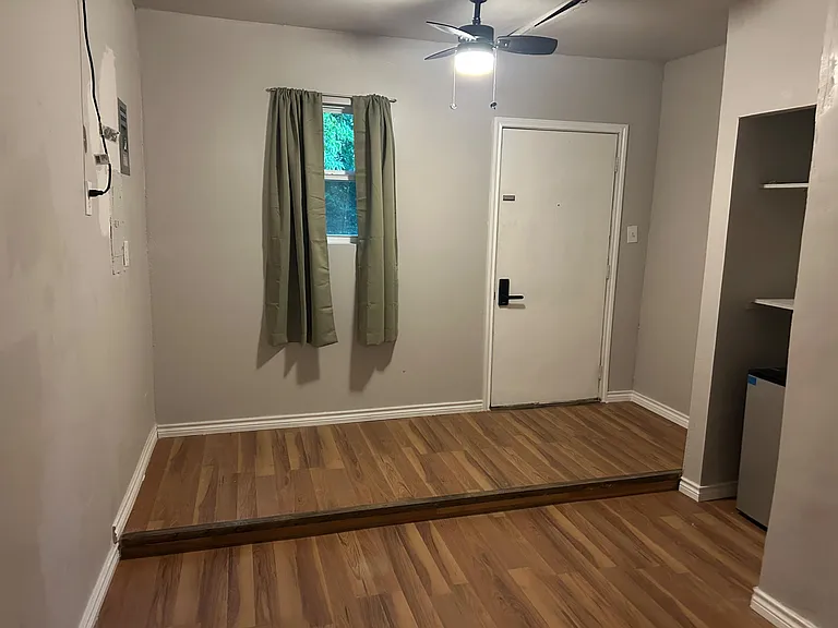 Open flex space with durable wood-look flooring, ceiling fan, a window with curtains, and built-in shelving for storage.