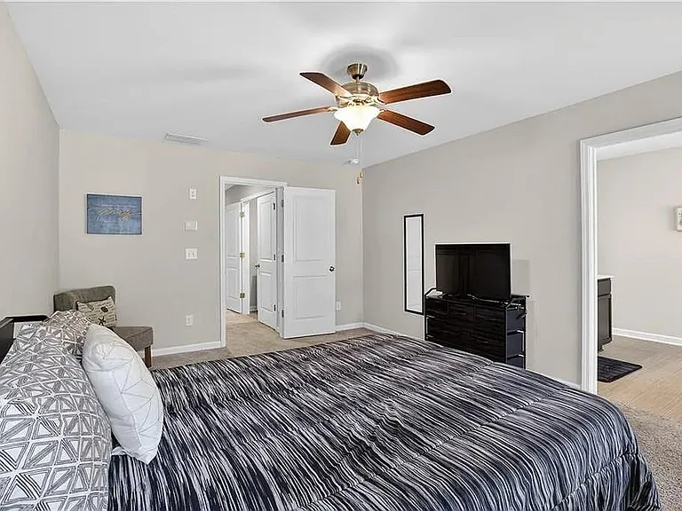 Spacious bedroom with ceiling fan, furnished bed, and dresser/TV setup, plus easy access to the adjoining hallway and bath.