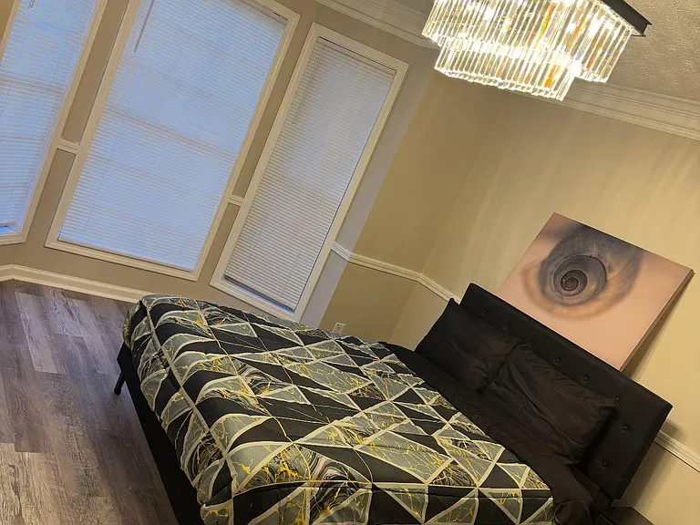 Bedroom with large windows, hardwood-style floors, and a furnished sleeping setup with bedside storage and overhead lighting.