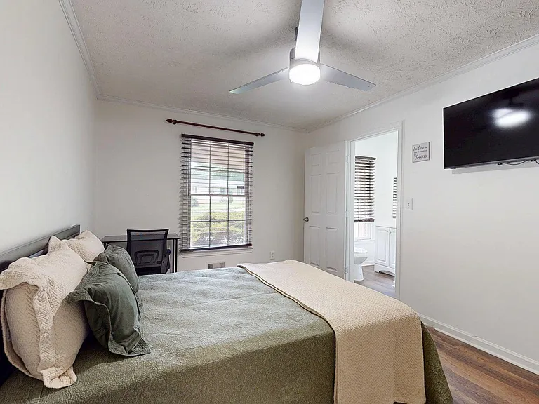 Furnished bedroom with a ceiling fan, wall-mounted TV, desk nook, and a window with blinds near the bath.