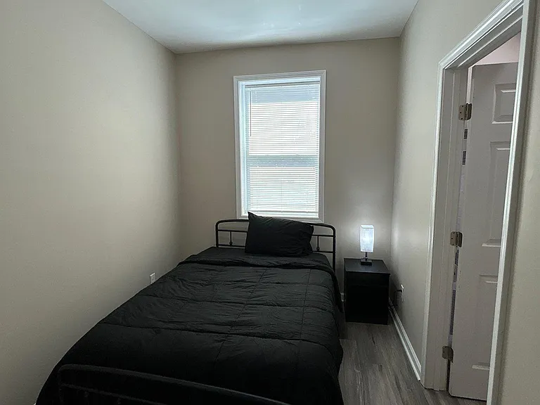 Private bedroom with a furnished bed, nightstand, and window with blinds, plus easy access to the nearby hallway.