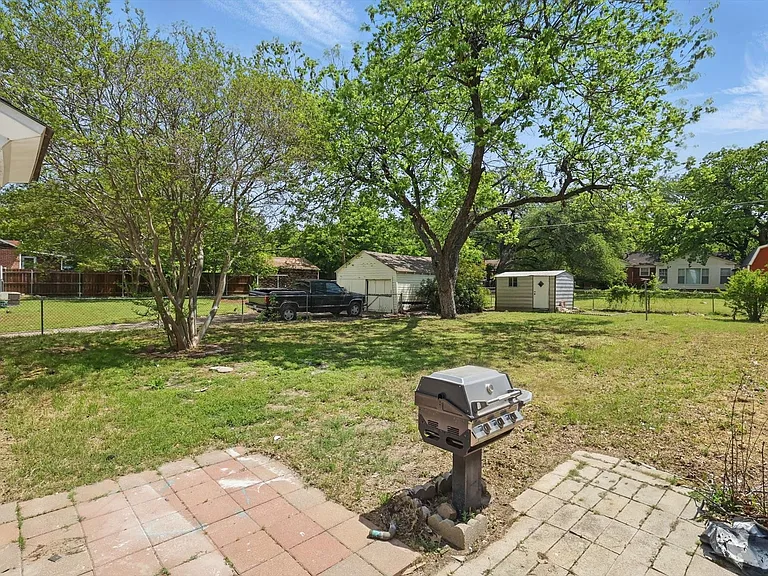 Expansive backyard with a paver patio, established trees providing natural shade, and a storage shed. Grill is non-functioning.