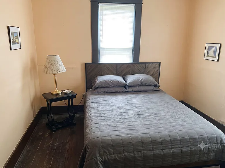 Furnished bedroom with a full bed, nightstand and lamp, plus a window with blinds for light and privacy.