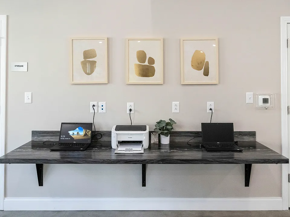 Modern home office setup with sleek black countertop, two laptops, a printer, and minimalistic wall art for a contemporary workspace.
