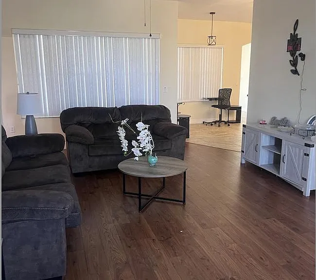 Open living area with vaulted ceiling, ceiling fan, wood-look floors, and large windows with vertical blinds for privacy.