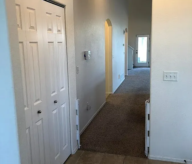 View of the foyer from the open wall of the office (no 4th wall or door). A coat closet, an alarm system on the wall to the left, followed by doorway to garage and powder room. The kitchen is to the right.