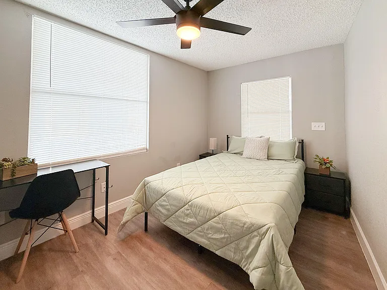 Furnished bedroom with a full bed, side tables, and a dedicated desk area, plus two windows and a ceiling fan.