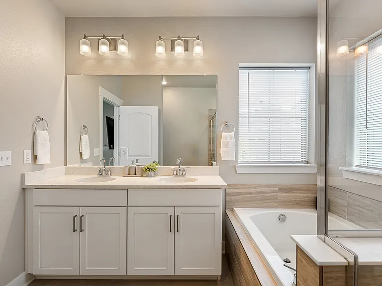 Modern bathroom with a double vanity featuring sleek white cabinets and chrome fixtures. Large mirror with bright lighting above, a spacious bathtub with wood-look tile surround, and a window with blinds provide a clean, bright, and inviting space.