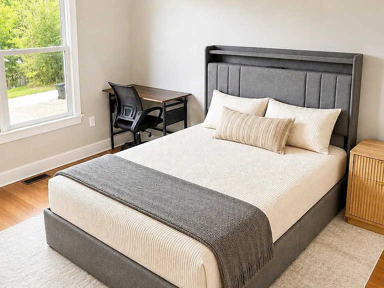 Furnished bedroom with a queen bed, bedside storage, and a dedicated desk area, plus a large window and ceiling fan.