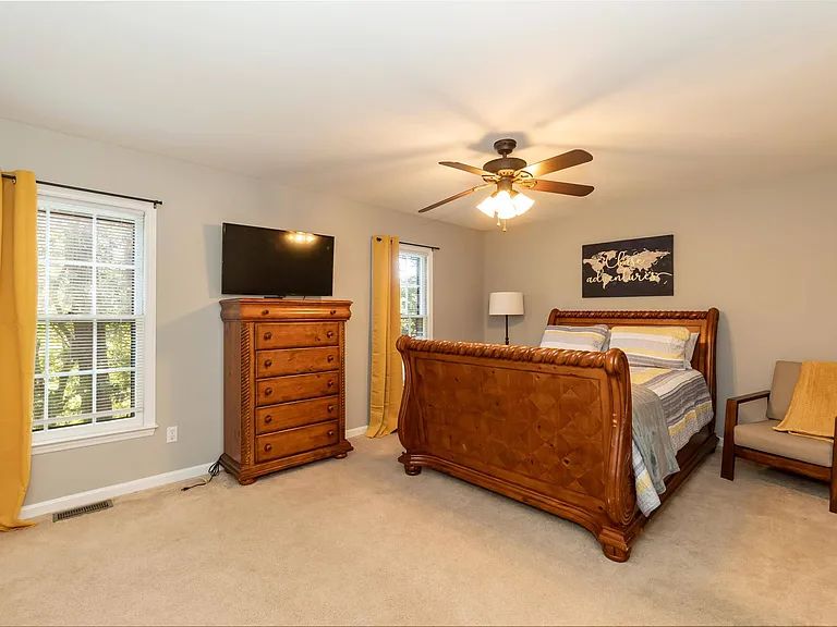Furnished bedroom with a wood bed frame, dresser, TV, ceiling fan, and two windows with curtains for natural light.