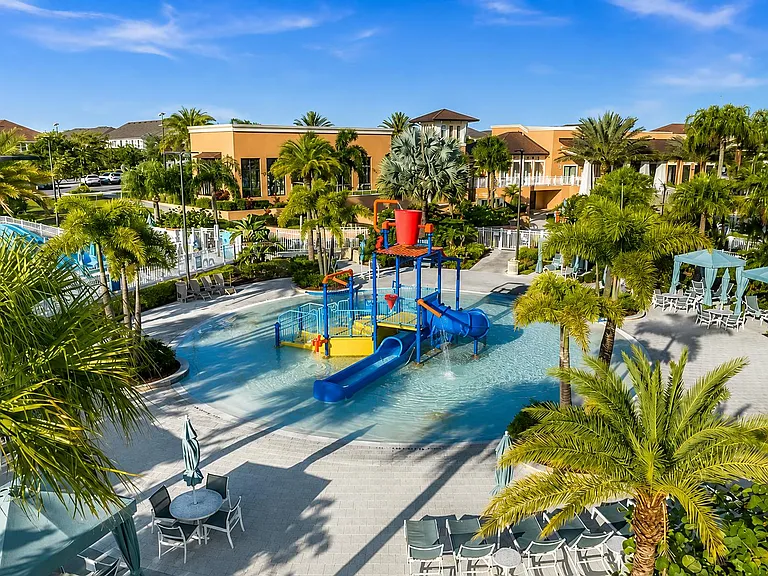 Resort-style outdoor pool area with splash pad and water slide, surrounded by seating, umbrellas, and tropical landscaping.