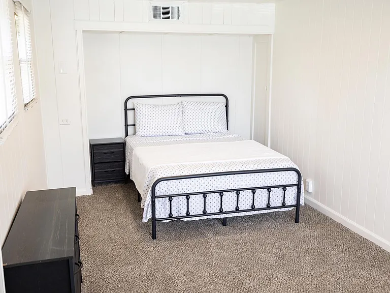 Private bedroom with a full-size bed, nightstand, and carpeted floors, plus a window with blinds for natural light.