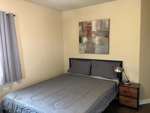 Neat bedroom featuring a full-size bed, nightstand with lamp, and a window with curtains for natural light.